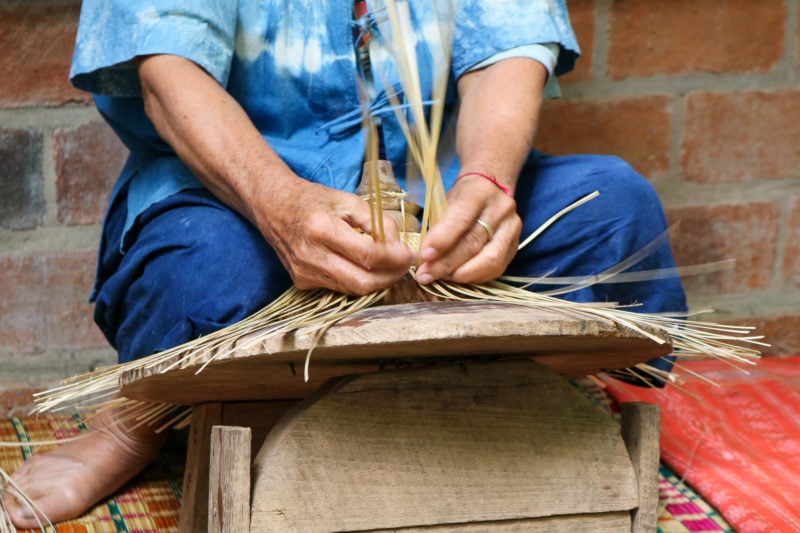 close up of an elderly lady's hands as she ties long pieces of dried grass into a conical shape to make a hat. the lady is wearing a blue tie-dyed t shirt and indigo-dyed trousers.