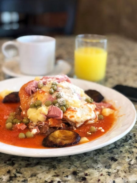 white plate on a table with a white coffee cup and a glass of orange juice behind, the plate has huevos motulenos on it, a fried egg on top of cripsy tortillas and covered with a thin tomato sauce, peas, pieces of pink ham, crumbled cheese, and pieces of fried plantain.