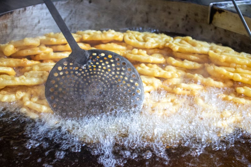 close up of a large black wok filled with hot bubbling oil and small crispy spirals of dough called jalebis with a large slotted spoon stirring them as they fry. Traditional Nepali desserts.