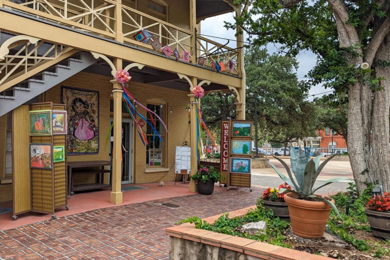 historic building with a collonaded veranda painted cream with stacks of paintings for sale outside in a stone courtyard in La Villita. What to do on a day trip in San Antonio Texas. 