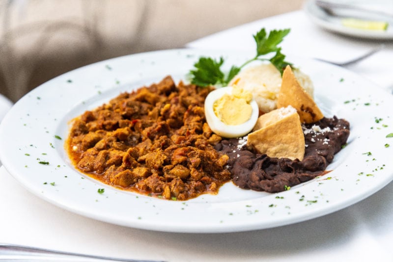 white plate with a brownish tomatoe sauce based stew on it filled with pieces of pork with two halves of a boiled egg on top and a small pile of brown refried beans next to the stew.