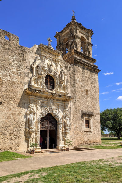 front of an old 18th century church built from beige coloured stone with a partly ruined front and carved facade. there is an oval shaped window above a large arched double door and a small square tower on the right side of the church. day trip in san antonio texas. 