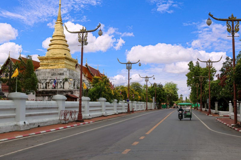 wide empty road in a city with a single tuktuk on it next to a white stone wall in front of a large golden stupa in the old city of Nan