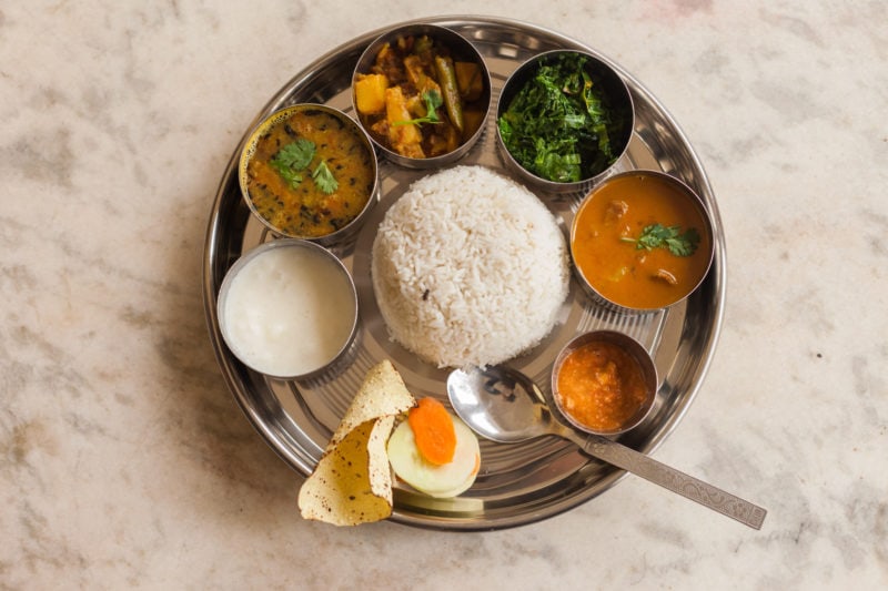flatlay of a silver thali plate with six small silver bowls and a piece of roti bread arranged in a circle around a mound of rice. The small bowls all contain different foods like curry, pickles, and leafy greens. 