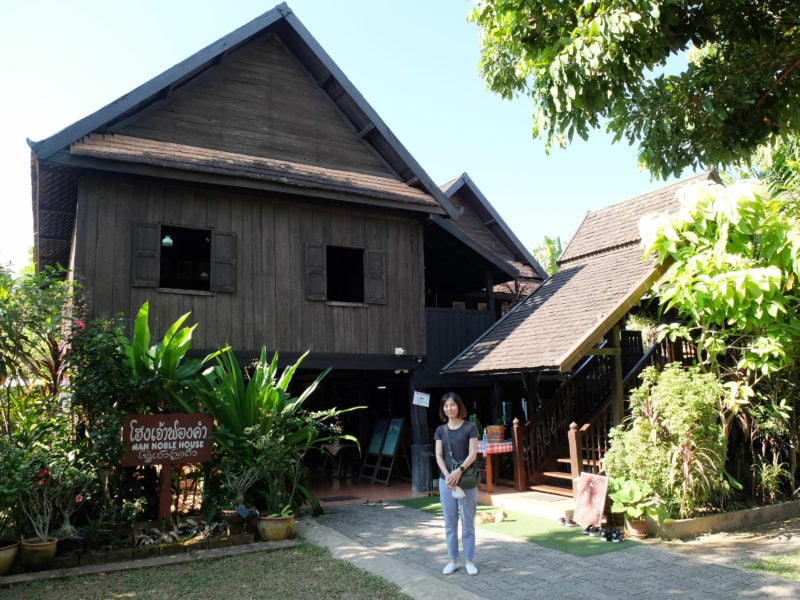 Thai woman in front of the Noble House, a large wooden two storey house build from dark teak wood and surrounded by green leafy bushes and trees in Nan province, Thailand.