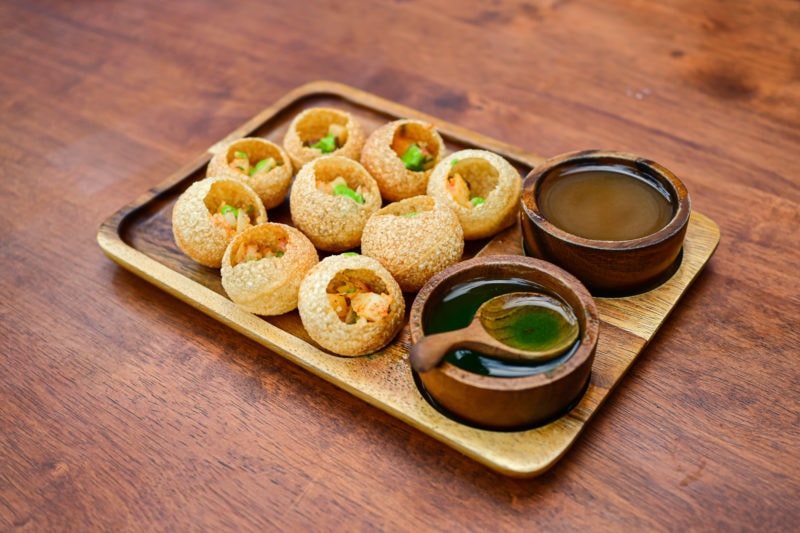 close up of a wooden tray on a wooden table with several small crispy dough balls on the tray with open tops and choped veg inside them. there are two small wooden bowls on the tray containing thin sauces and a wooden spoon. Pani Puri is a popular street food in Nepal 