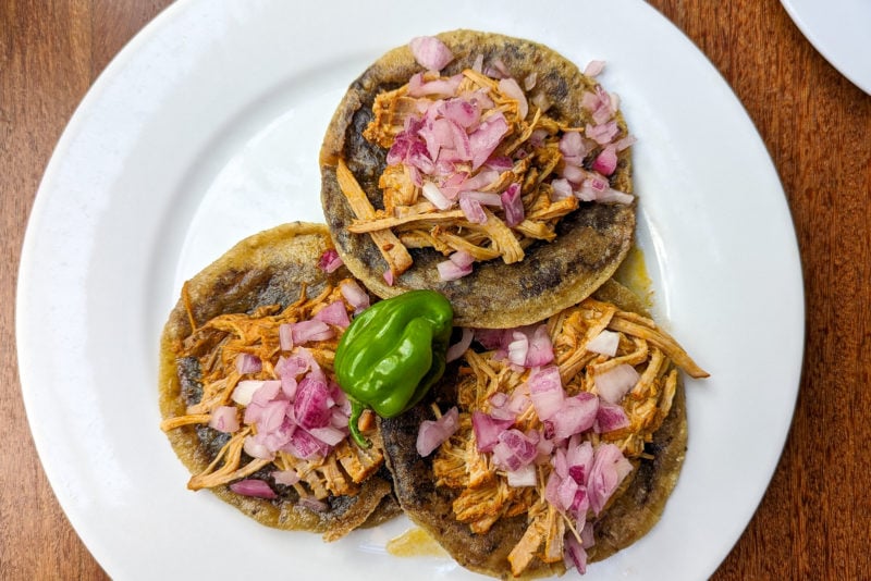 flat lay of three small fried torillas on a white plate, each topped with shredded pork in a reddish brown sauch called cochinita pibil and pink pickled onions with a single green habanero in the middle of the three tortillas