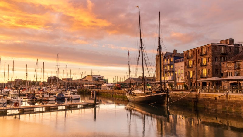 a large harbour filled with sailing boats reflected in the very calm water with a few histroic stone buildings behind at sunset with an orange sky overhead