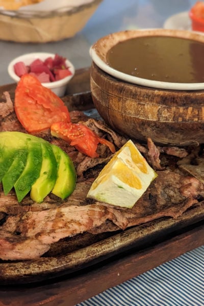 wooden platter with grilled brown pork, a slice of peeled yellow orange, chopped slices of avacado arranged in a fan, pieces of cooked tomato, and a wooden bowl filled with a brown soup made from beans.