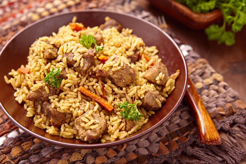 Close up of a brown china bowl on a woven placemat filled with yellow rice pilaf with lamb meat, sliced carrots, and pieces of fresh green herb