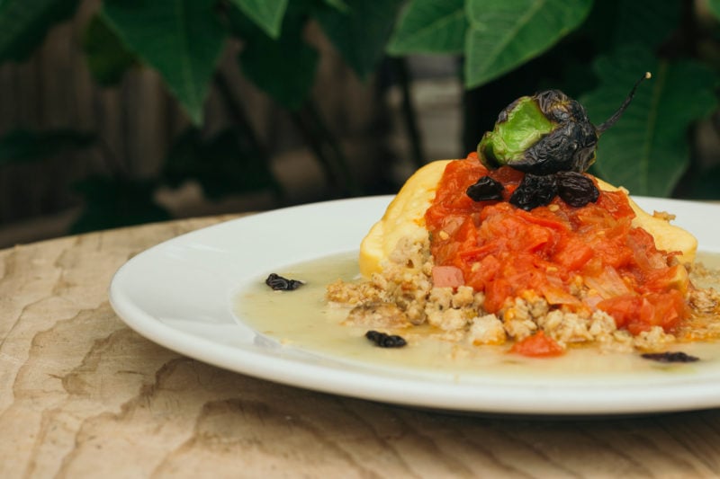 white plate on a wooden table with a yucatecan queso relleno on it, a piece of cheese covered with a tomato sauce and surrounded by a creamy sauce