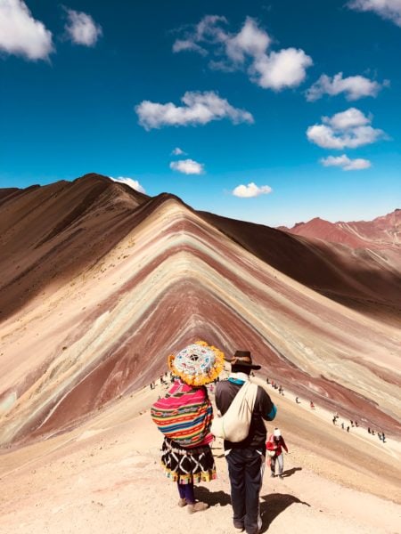 rainbow mountain in peru, a man and woman in traditional dress walking up the ridge of the mountain which is striped in different shades of red brown and beige on a bright sunny day with blue sky overhead. the man is wearing a black jacket and black cowboy hat, the woman is carrying a large multicoloured sack over her shoulder and a colourful parasol with a yellow fringe.