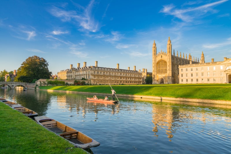 view of the River Cam in Cambridge with a single wooden punting boat on the still river water. there is a large beige coloured stone building on the other side of the river and its towers are reflected in the water. 