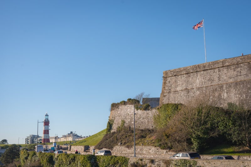 grey stone fort with a partly ruined wall in two tiers against a blue sky with a red and white lighthouse in the distance