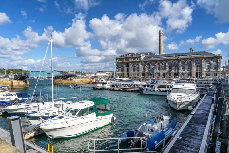 small marina with several yachts on bright blue water with a large grey stone building behind