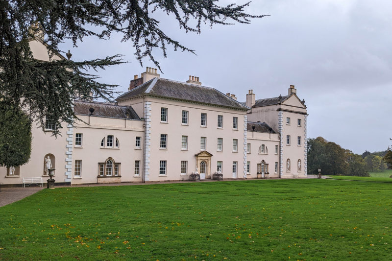 large cream painted english country manor house with a grassy lawn in front and grey sky behind