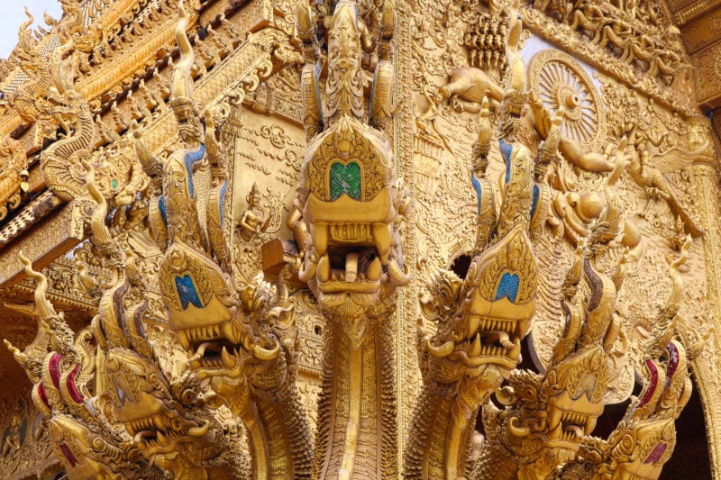 close up of a section of a statue of a dragon like creature with several heads all carved from gold with a golden temple behind