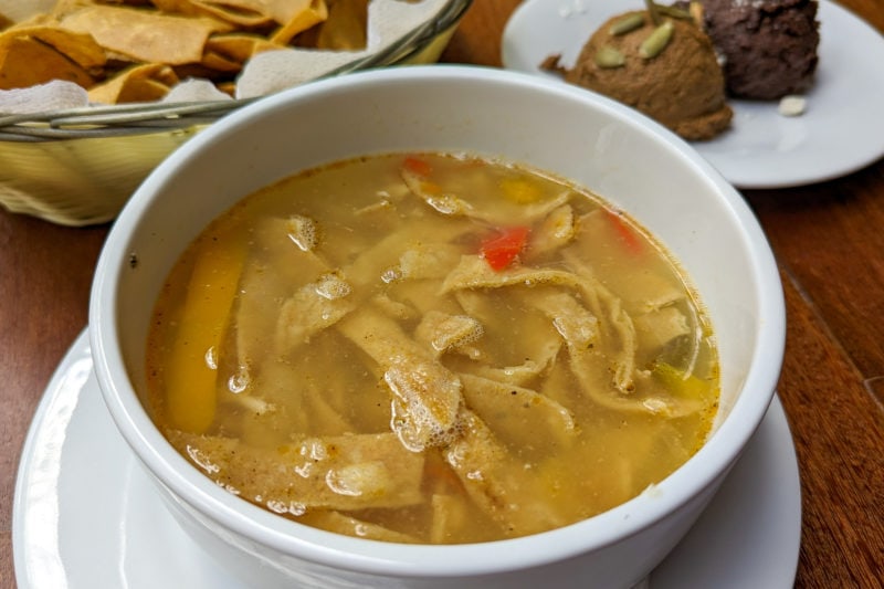 close up of a white china bowl filled with yellowish brown soup with shredded chicken and broken pieces of fried tortilla crisps in it