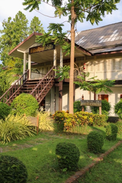 outside of a large two storey mansion built from white painted teak wood with an unpainted wooden staircase leading up to a large wooden porch on the upper floor and a grey tiled roof above