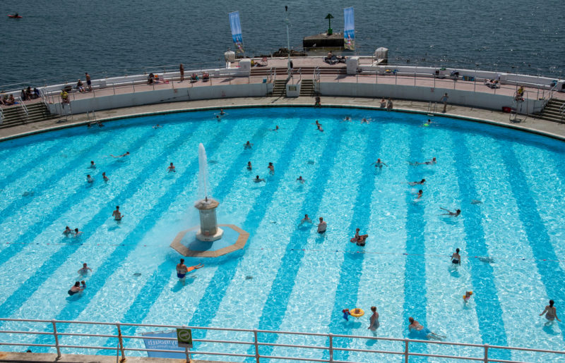looking down at a large semi circular outdoor swimming pool lido with a stone wall around it and the sea beyong in Plymouth England