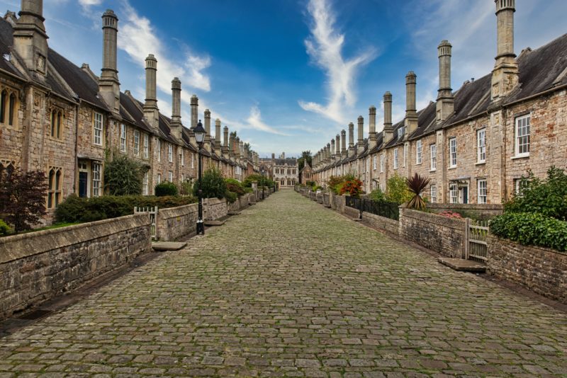 grey stone cobbled street lined with matching terraced brownish-grey stone cottages with dark grey tiled roofs running on either side of the street with a blue sky overhead. 