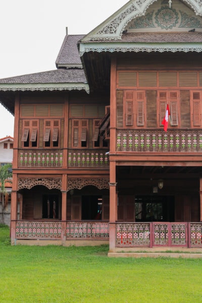 exterior of a two storey mansion built from reddish coloured teak wood with a grey tiled triangular roof. the second floor is lined with rows of narrow shuttered windows and there is a verandah around the ground floor of the property.  things to do in phrae thailand.