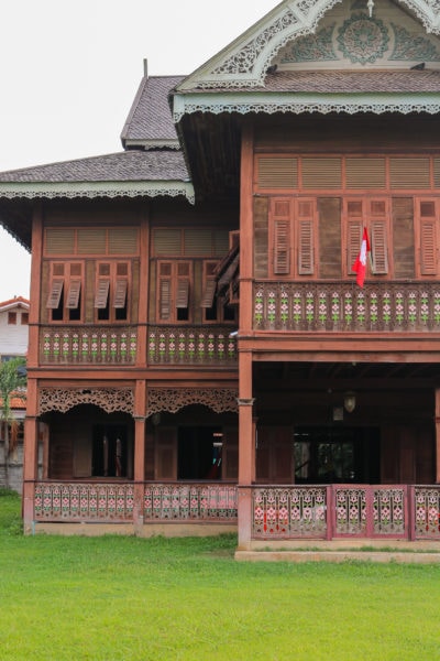 exterior of a two storey mansion built from reddish coloured teak wood with a grey tiled triangular roof. the second floor is lined with rows of narrow shuttered windows and there is a verandah around the ground floor of the property. things to do in phrae thailand.