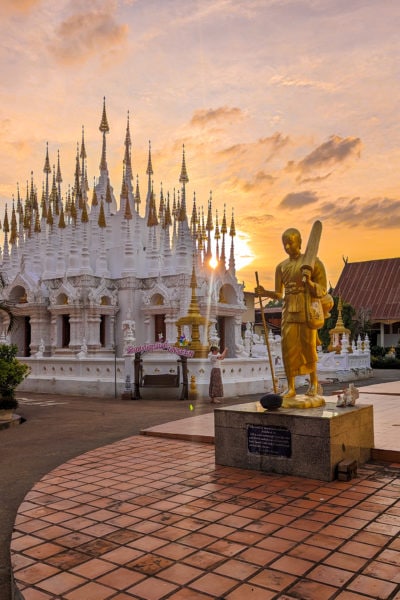 courtyard of a temple in thailand with a small white building covered in small conical turrets with golden pointed roofs. there is a golden statue of a man holding a staff in front on a red tiled floor and the sun is setting behind the white building with lens flare between the turrets.  things to do in phrae thailand.