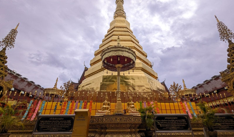 tall golden stupa at a thai temple with a small golden umbrella-like statue in front and a long golden fence around it wrapped in orange fabric. it is a grey day with thick clouds behind the stupa.