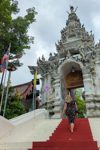 emily in a long blue dress with flowers walking up the top of an outdoor staircase with white stone steps with a red line down the middle going uphill to a carved stone archway at a temple in thailand