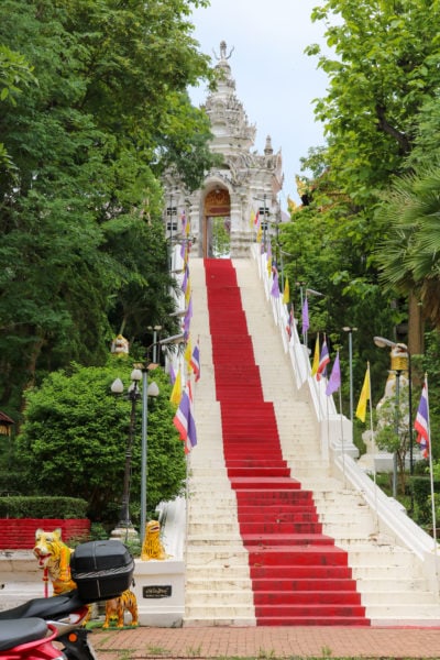 tall outdoor staircase with white stone steps with a red line down the middle going uphill to a carved stone archway at a temple in thailand with trees on either side of the staircase.  things to do in phrae thailand.