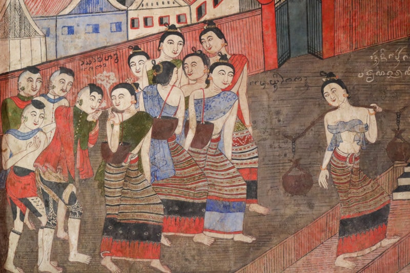 close up of a section of an old mural with a mostly red background showing a group of women in traditional Thai dress whispering and talking to each other in front of a wall with part of a city visible behind