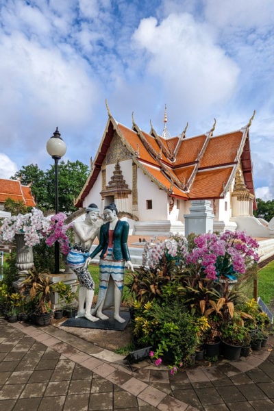 white thai buddhist temple with red tiled roof. in front is a statue of two lovers with the man whispering to the woman. the statue is surrounded by pink flowers