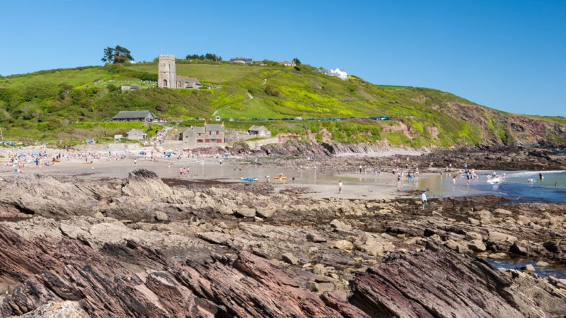 small shingle beach with rocky shelves and a low green cliff behind on a sunny day