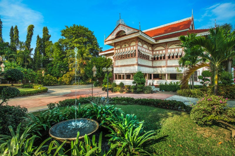 The Wongburi House In phrae Province a large white wooden mansion with wooden shutters and pink details with a large garden in fornt with trees and a pond and a small fountain