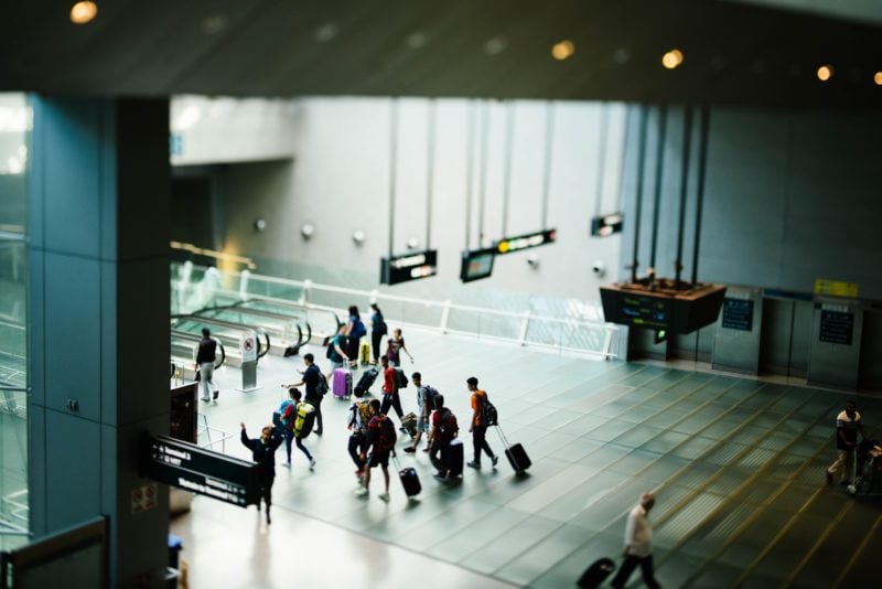 aerial shot of a waiting room in an airport with a crowd of people walking through, many pulling wheeled suitcase, the floor and walls are light grey and there are signs hanging down from the cieling