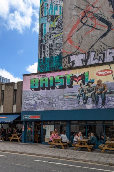 street scene in Bristol England with a large mural on a billboard above a blue painted cafe with outside seating. the mural shows a group of men sitting on a girder over a city skyline and says love from Bristol in big green letters. There is part of another street art mural showing on a grey wall on the side of a tower block above this. 