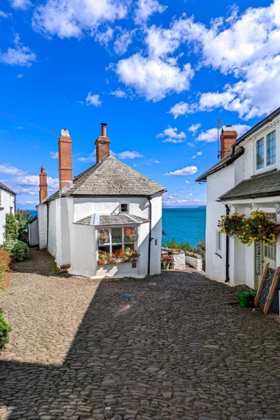 cobbled street with two white cottages on either side at the end and the blue sea visible between them, on a very sunny day with blue sky above, both cottages have grey slate roofs and red brick chimneys and large hanging baskets full of flowers above the windows