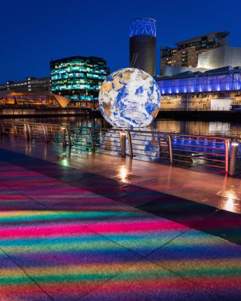 Salfrod Quays in Manchester at night with a huge lit-up model globe floating in the river next to a stone walkway with multicoloured lights projected onto it in a line. behind are several buildings and skyscrapers lit up at night. 