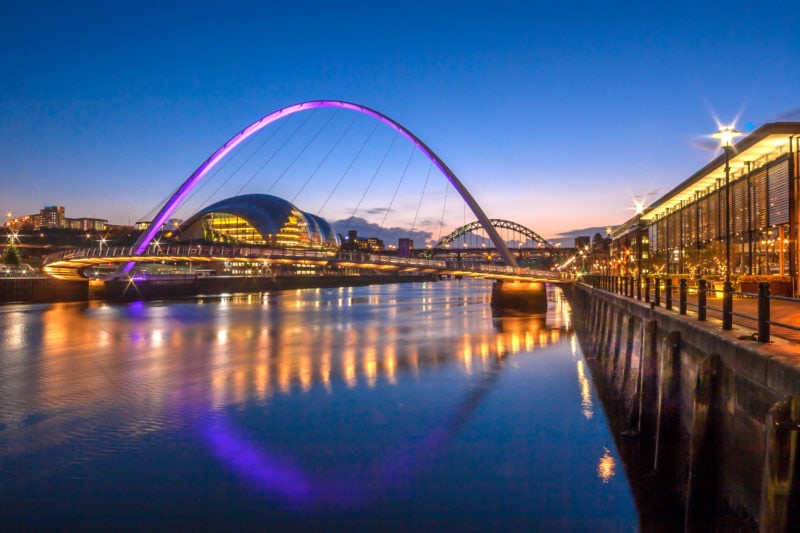The Gateshead Millennium Bridge, a tilting pedestrian bridge spanning the River Tyne in NE England, taken at night with the tall arch of the bridge lit in bright magenta against a clear dark blue sky. The bridge links Gateshead's Quays and the Newcastle upon Tyne Quayside. Among the buildings are the  Sage Music Centre, and The Tyne Bridge. All the buildings are lit up at night and reflected in the still water of the river. 