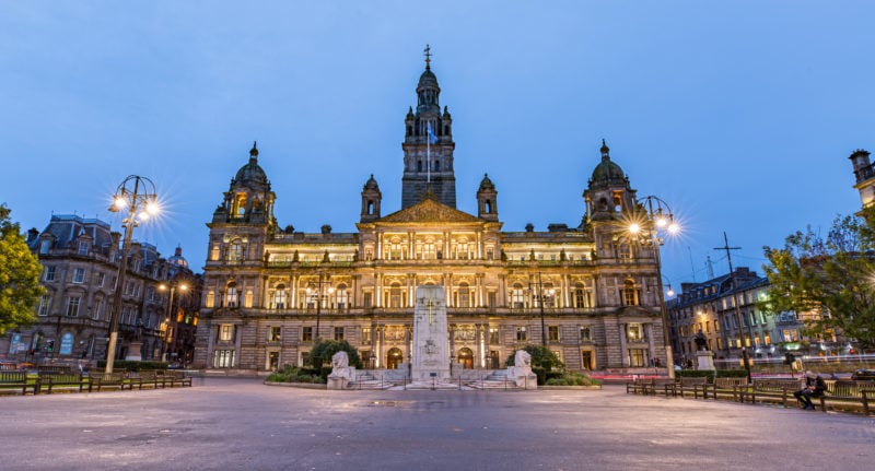 large historic stone palace style builing in Glasgow's Goerge Square at night with the building lit up in yellow against the clear blue sky and the square in front almost empty.