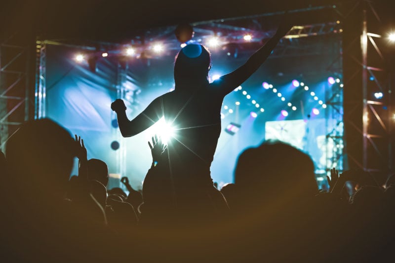 Girl silhouette on someone's shoulders above a crowd of people in a nightclub with her hands raised hands in front of a stage lit up in blue at a music concert. Best cities in the UK for nightlife. 