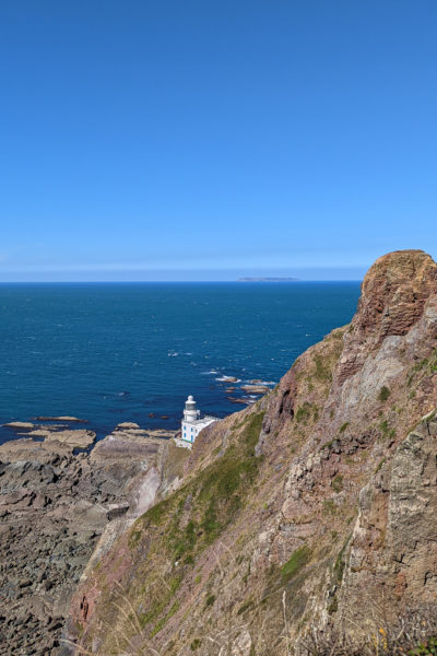 view from the top of a peninsula with a grey-brown rocky cliff falling away to a small white lighthouse on the tip and the blue sea beyond that on a very sunny day with clear blue sky above