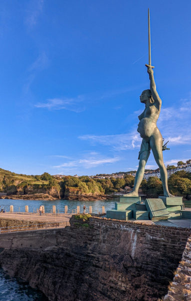 grey stone harbour wall with a very tall greenish metal statue of a naked pregnant woman holding a sword above her head, the blue water of th eharbour is behind her and a tree covered hill on the far side of that, on a very sunny day with blue sky above.