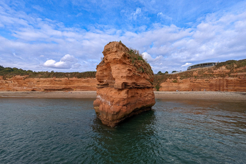 view of a bay from the sea with a large red coloured rock stack in the middle of the bay and low red cliffs framing the shingle beach on a sunny day with blue sky above