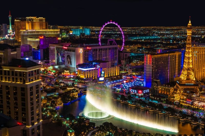 aerial view of the Las Vegas skyline at night with a long vountain lit up white and many buildings and skyscrapers all lit up in different colours. 