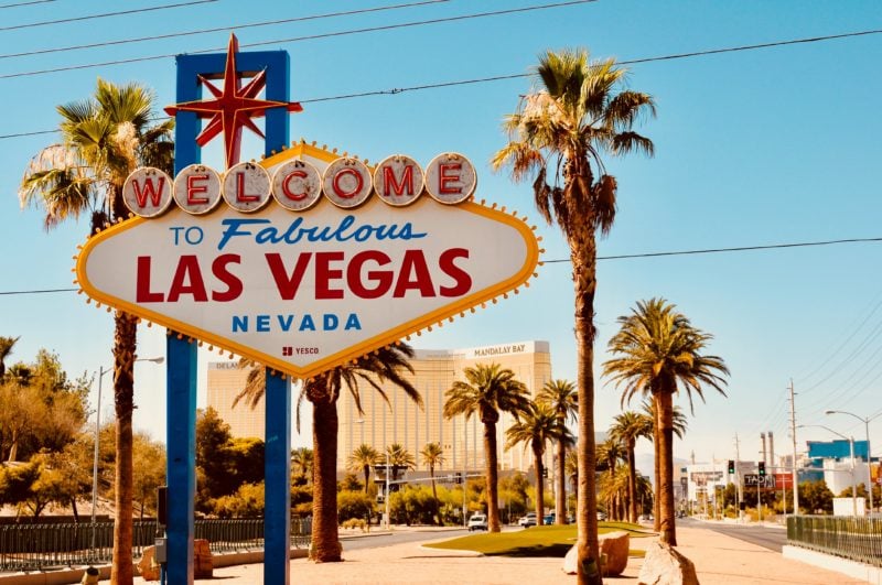 street sign with neon lettering saying welcome to fabulous las vegas nevada next to a road lined with tall palm trees on a sunny day with blue sky above