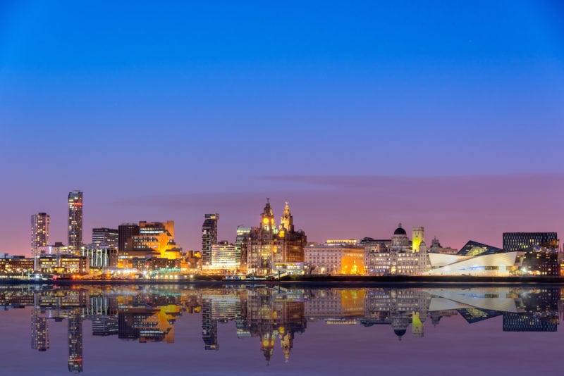 The Liverpool skyline at night from across Mersey river with all the major buildings such as liverpool museum, three graces, Royal Liver building, Port of Liverpool etc, all lit up against a clear blue evening sky and reflected in the still water of the river. best uk cities for nightlife. 