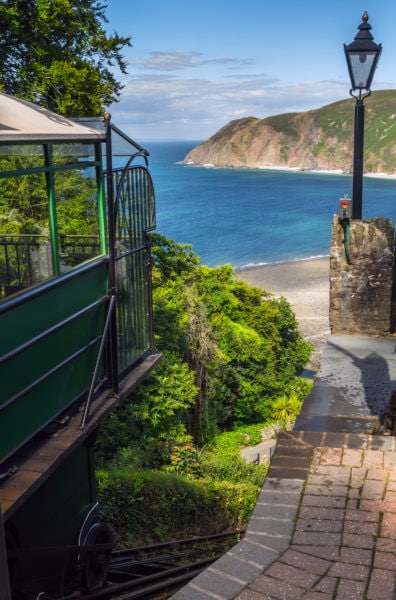 edge of a dark green cliff railway carriage at the top of a cliff with a view of the blue sea below and some low grass topped cliffs across the bay
