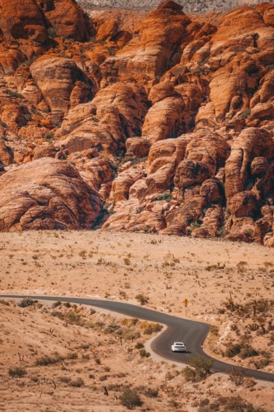 winding road with a single white car in Red Rock Canyon park with desert sand on either side of the road and a large orange rock cliff face on the far side